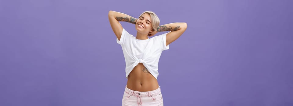 Great feelings after sunbathing. Chill and relaxed attractive young female in cropped top with perfect body and tattoos holding hands behind head, stretching with delight and smiling with closed eyes.