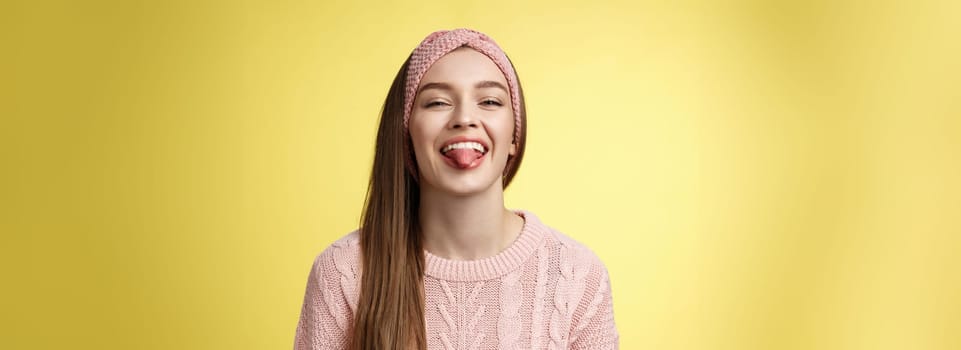 Positive entertained cute funny glamour young european girl in sweater, knitted trendy headband smiling fooling around showing tongue playfully, mocking friend enjoying sunny day over yellow wall.