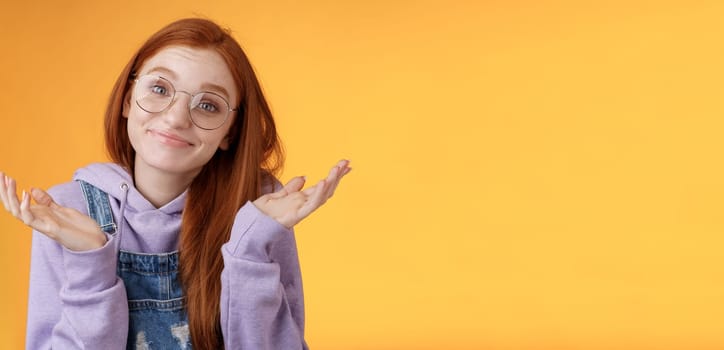 Nothing much. Attractive silly unsure carefree redhead unbothered cute female wearing geek glasses smirking uncertain shrugging hands spread sidways clueless what do future, orange background.