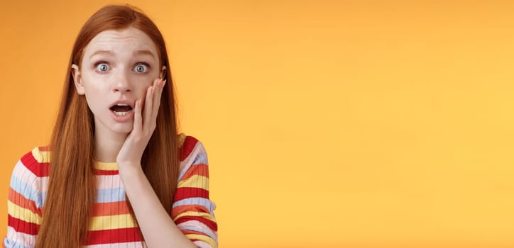 Concerned worried ginger girl blue eyes drop jaw gasping touch cheek confused looking nervously anxious, show empathy hearing terrible upsetting story standing orange background. Copy space