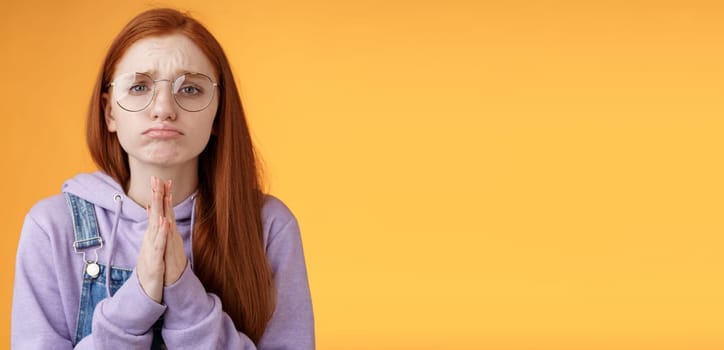 Please beg you. Clingy upset sad caucasian redhead girlfriend wearing glasses frowning grimacing pouting pleading help asking favour need urgently borrow something, orange background.