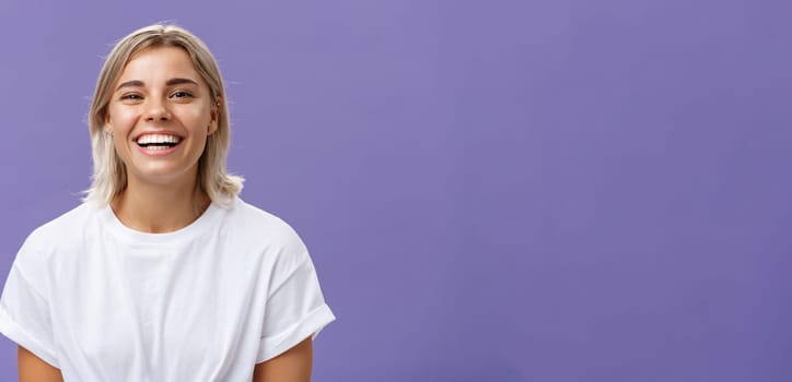 Close-up shot of joyful charming blonde female with delighted and pleased smile standing in white t-shirt over purple background spending time in awesome amusing company. Emotions concept