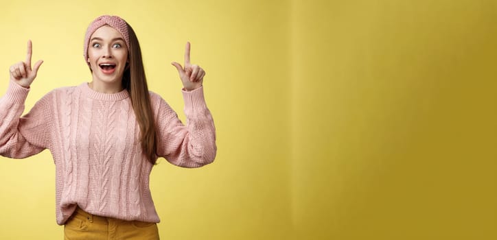 Ecstatic amused positive young 20s woman wearing casual sweater, headband dropping jaw amazed looking astonished, surprised pointing up excited, upbeat of seeing incredible promo over yellow wall.