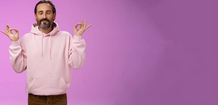 Peaceful charming hipster old man wearing cool pink hoodie close eyes breathing practice release stress meditating standing relaxed happy yoga meditation pose nirvana gesture, purple background.