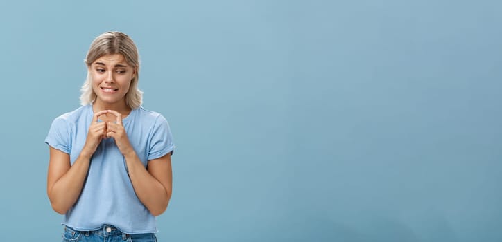 Lifestyle. Studio shot of insecure and timid silly girlfriend with blond hair clenching teeth looking shy down and steepling index fingers unconfident trying ask out boy she likes over blue background.