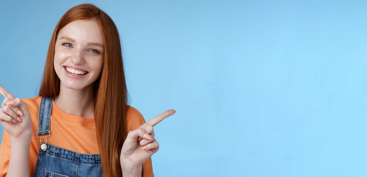 Friendly happy laughing pretty redhead girl female student showing lots opportunities advice make choice pointing sideways left right introducing different product grinning gladly, blue background.