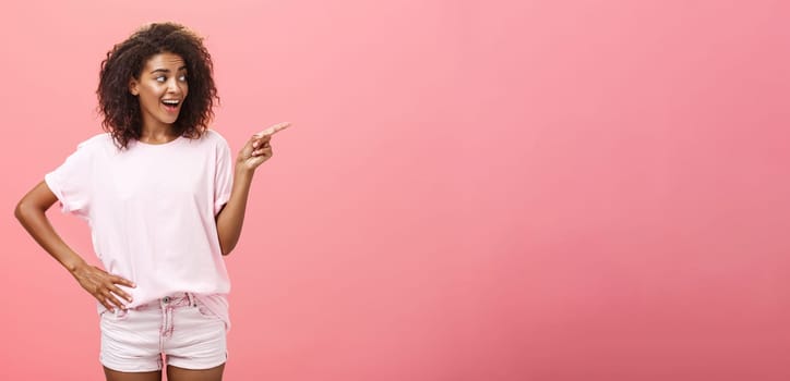 Hey wanna go there. Portrait of intrigued pleased good-looking stylish african woman on vacation wearing summer clothes pointing and gazing right with curious look over pink background. Advertisement concept