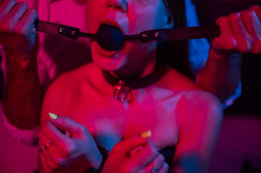 A man gags a woman in a pink blue neon light in the bedroom. BDSM concept