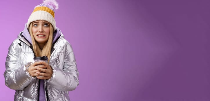 Shaking uncomfortable displeased girl wearing stylish silver jacket hat trembling freezing cold warm hands holding take-away coffee cup clench teeth pop eyes discomfort, standing purple background.