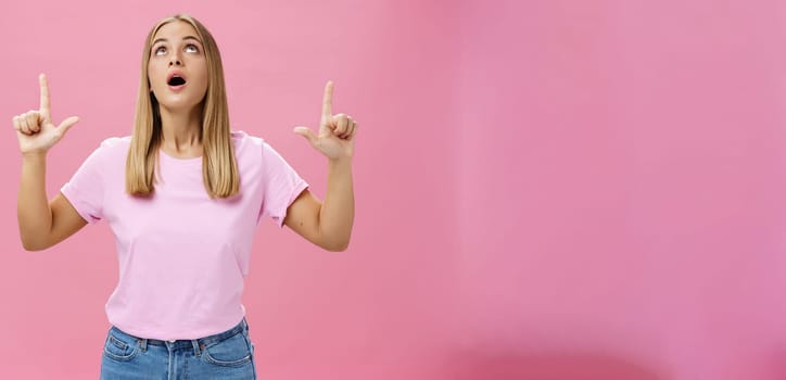 Portrait of amused and impressed curious attractive woman in t-shirt dropping jaw looking and pointing up astonished and intrigued watching interesting object in sky posing against pink background. Lifestyle.