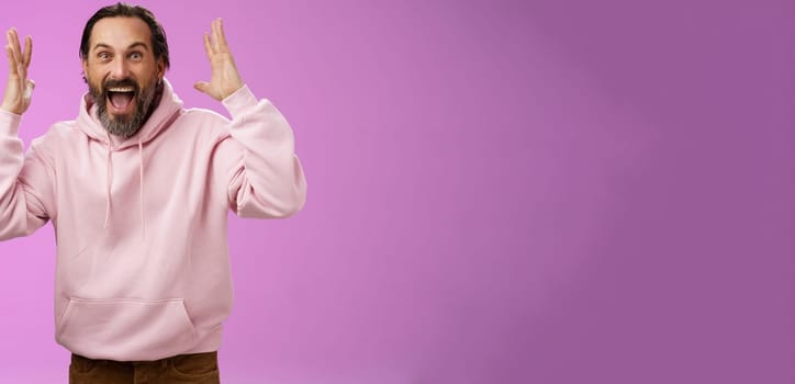 Happy excited lucky adult bearded man celebrating excellent news raising hands gesturing thrilled smiling broaldy triumphing victory win, yelling gladly achieve goal, standing purple background.