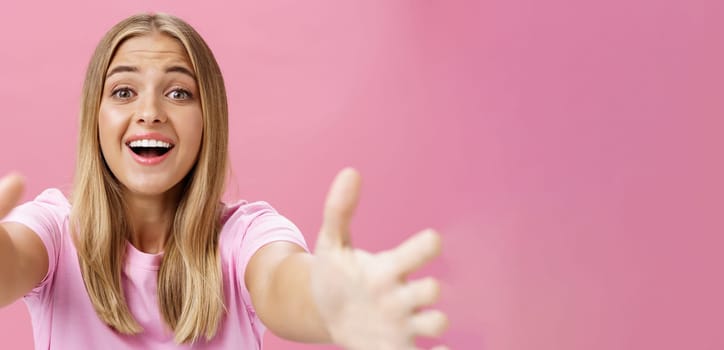 Close-up shot of caring and loving silly girl with tanned skin and fair hair pulling hands towards camera to give warm hug smiling broadly gazing with admiration wanting cuddle over pink background. Lifestyle.