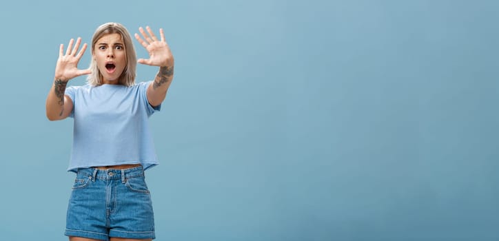 Woman terrified screaming and asking stop. Portrait of shocked panicking troubled blonde female in denim shorts and casual t-shirt pulling hands in no gesture dropping jaw and frowning. Copy space