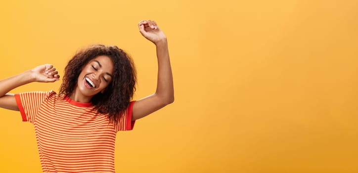 Waist-up shot of relaxed and relieved happy african american young female student in striped t-shirt stretching arms up closing eyes from delight and smiling having great nap over orange background. Lifestyle.