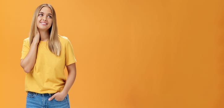 Pretty and tender cute european female independent freelancer in yellow t-shirt sighing touching neck and gazing dreamy at upper right corner with pleasant smile, posing over orange background.