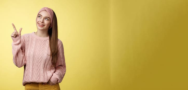 Portrait of amused interested cute 20s european girl wearing sweater, headband looking upper left corner pointing sideways smiling intrigued, ecstatic, captured by curious promo over yellow wall.