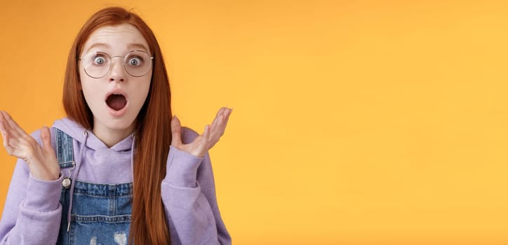 Close-up shocked sensitive concerned young panicking redhead woman worry drop jaw gasping raise hands spread freak out stare surprised emotional reacting incredible news, orange background.