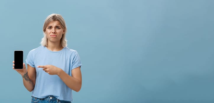 Lifestyle. Unhappy sad attractive silly girl with fair hair tattoos and tanned skin frowning with regret and disappointment poonting at smartphone screen as if being dissatisfied with new girlfriend of ex-lover.