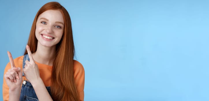 Lifestyle. Happy tender redhead girl sincerely smiling white teeth helpful look camera excited give hand pointing upper left corner introduce sale offer recommend try promo standing blue background.