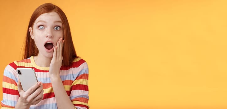 Concerned shocked emotive redhead girl find out stunning news look camera drop jaw gasping impressed touch cheek hold smartphone finish reading impressive disturbing story, orange background.