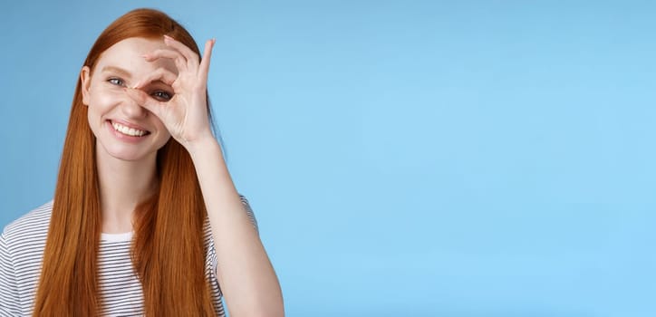 Charismatic happy adorable redhead teenage girl sincere eyes making circle eye show okay ok sign delighted like approve cool idea smiling satisfied achieve perfect score, standing blue background.