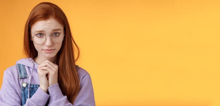 Silly guilty young shy redhead girlfriend asking forgiveness supplicating lower head look from under forehead frowning begging apology favour standing insecure sad pleading help, orange background.