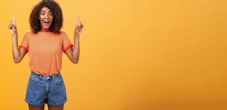 Charmed and impressed african american stylish woman seeing incredible and awesome item on sale raising hands and pointing up with broad grin being excited of new purchase over orange wall. Emotions and advertisement concept