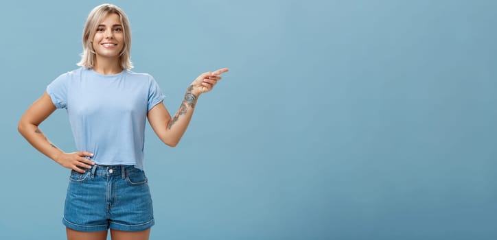 Check it out. Portrait of beautiful confident young caucasian girl with blond hair pointing right and holding hand on waist in self-assured relaxed pose smiling broadly over blue background.