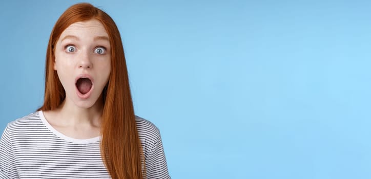Wow so cool. Impressed speechless amused attractive redhead girl blue eyes freckles open mouth wide omg drop jaw astonished express surprise amazement, standing blue background. Copy space