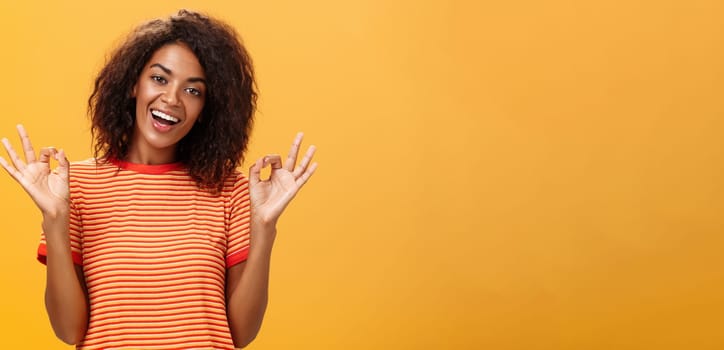 Waist-up shot of chill outgoing calm girlfriend with confident look in trendy striped t-shirt showing okay or excellent gestur and smiling broadly assuring everything alright over orange background. Lifestyle.