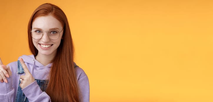 Confident good-looking sassy smiling redhead freelancer girl wearing glasses pointing upper left corner index fingers grinning white teeth assertive suggest recommend promo, orange background.
