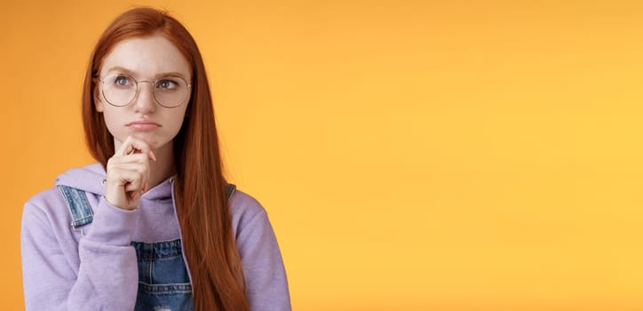 Puzzled serious-looking smart young redhead girl confused thinking deep focus problem touch chin frowning look upper left corner thoughtful, pondering suspicious situation, orange background.