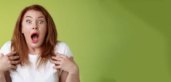 Shocked panicking redhead middle-aged woman gasping drop jaw open mouth stare camera freak-out anxious pointing herself impressed terrified frustrated nervously react green background.