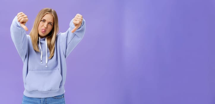 Indoor shot of moodie displeased and unimpressed cool modern female with blond hair and tanned skin tilting head sticking out tongue showing thumbs down in dissatisfaction posing against purple wall. Body language concept