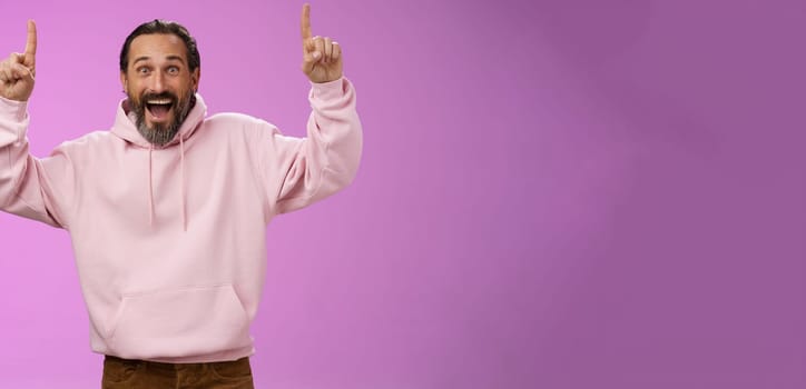 Energized happy good-looking trendy mature dad beard grey hair in pink stylish hoodie wearing son outfit pointing up index fingers thrilled favorite music group in town, standing purple background.