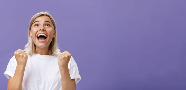 Waist-up shot of grateful delighted lucky girl with attractive tan in white t-shirt clenching fist from positive emotions gazing delighted and happy up thankful giving thanks god for fulfilling dream. Emotions concept