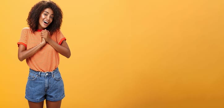 Girl sighing and gazing at cute thing clenching palms together near heart smiling with admiration and love being feminine and tender posing over orange background feeling affection. Copy space