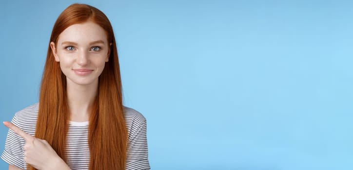 Attractive modest good-looking redhead girlfriend acting mature friendly smiling pointing left index finger giving direction show way bathroom standing blue background joyful kind grin.