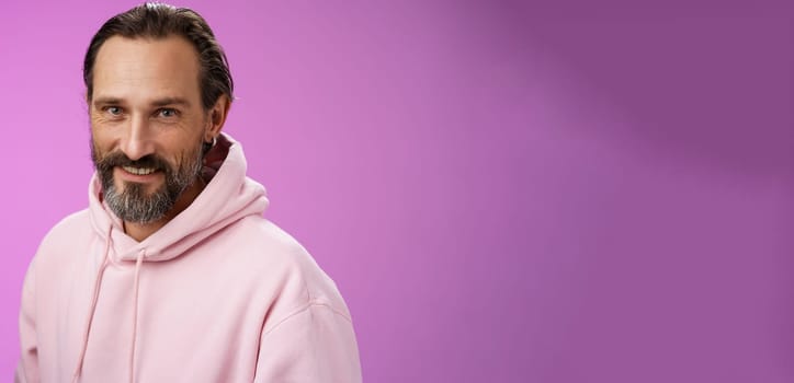 Charming alluring handsome bearded stylish adult male model earring pink hoodie smiling delighted express confidence positivity feel lucky amused, standing purple background talking casually.