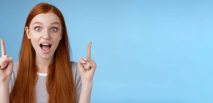 Surprised happy enthusiastic young redhead woman reacting impressed upper hanging promo pointing up index fingers drop jaw amused look thrilled camera telling about awesome offer, blue background.