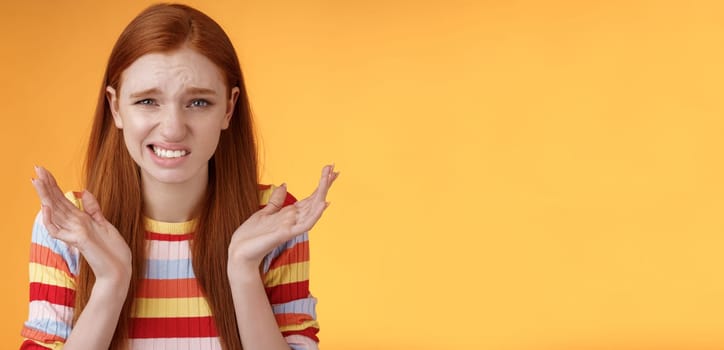 Awkward unhappy worried young redhead girl cringe feel sorry apologizing smirking smiling nervously frowning squinting spread hands sideways shrugging confused, standing orange background.