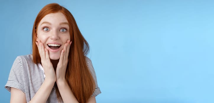 Amused happy dreamy young excited redhead female fan looking fascinated camera smiling admiration delighted touch face thrilled express emotional cheerful surprise, blue background.