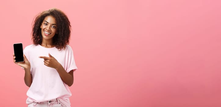 Suggest this model of phone. Happy charming african american curly-haired woman in stylish outfit holding smarpthone and pointing at device screen while recommending cool app over pink wall.