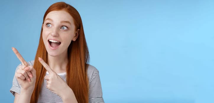 Lifestyle. Amused adorable redhead european girl say wow stare gasping astonished pointing looking upper left corner excited smiling drop jaw amazed standing thrilled blue background excitement concept.