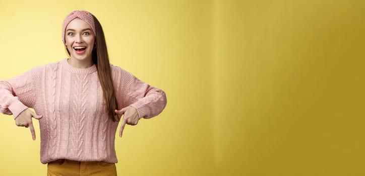 Lifestyle. Euphoric attractive young trendy girl in knitted band wearing sweater grinning excited enthusiastic pointing down thrilled of awesome promotion standing amused and overwhelmed over yellow background.