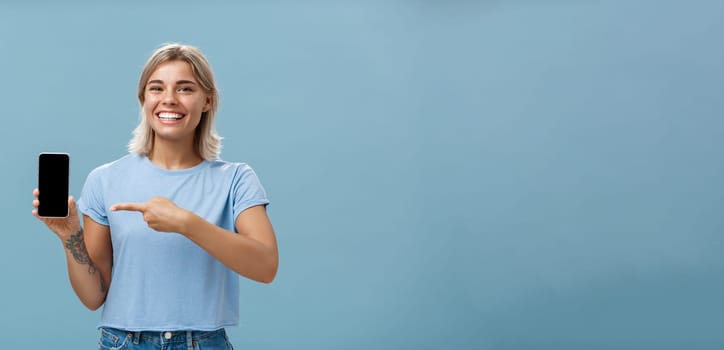 Cute enrgized girl showing smartphone. Charming blonde young female with cool tattoos laughing and smiling from happiness and joy pointing at device holding phone with screen faced to camera.