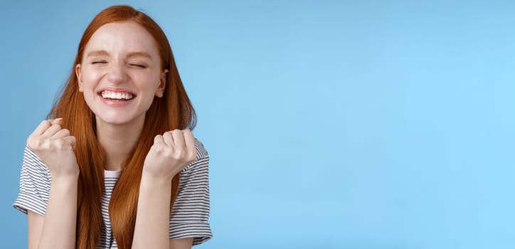 Lifestyle. Happy accomplished good-looking ginger girl receive positive results test clench fists rejoicing yelling yes celebrating do winner dance close eyes relieved standing blue background triumphing.