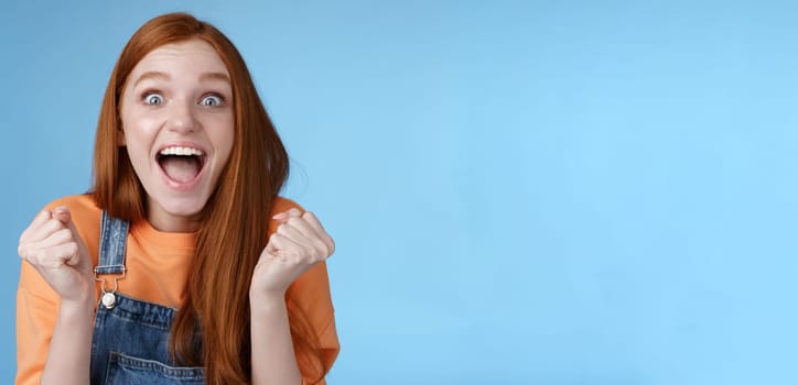 Lifestyle. Excited happy overwhelmed cute ginger girl blue eyes yelling out loud rejoicing fantastic awesome news clench fists triumphing celebrating victory win standing amazed blue background achieve success.