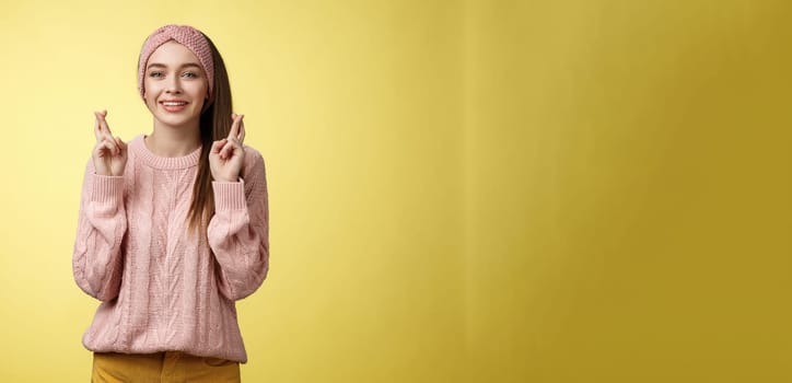 Cross my fingers for you. Supportive cute young european girlfriend wearing sweater, glamour headband making luck, wish gesture smiling hopeful having faith dream come true over yellow background.