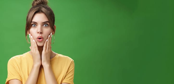 Portrait of impressed and shocked cute feminine woman in yellow t-shirt folding lips from excitement and interest touching cheeks surprised reacting to amazing rumor posing over green background. Body language concept